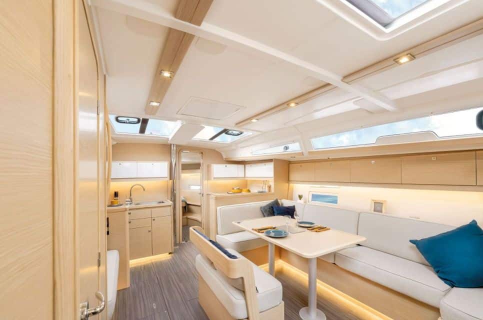 Interior del velero Amodiño iluminado con luz natural y luces LED, espacio tipo salón con sofá y mesa, pequeña cocina y camarote al fondo